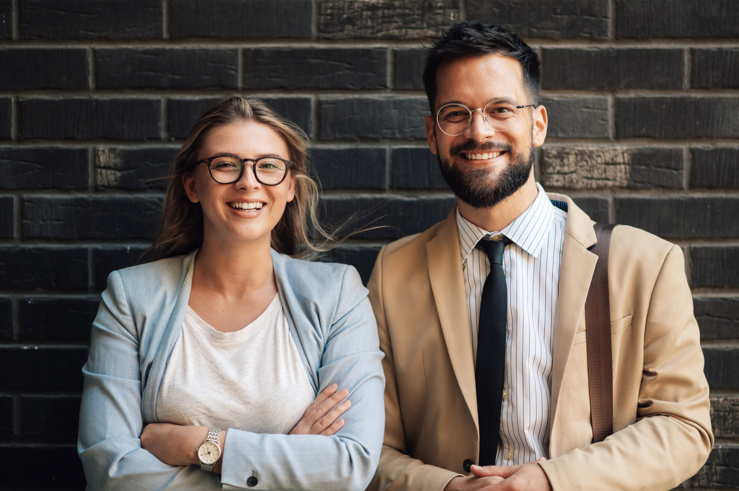 two young professionals smiling with arms crossed against brick wall